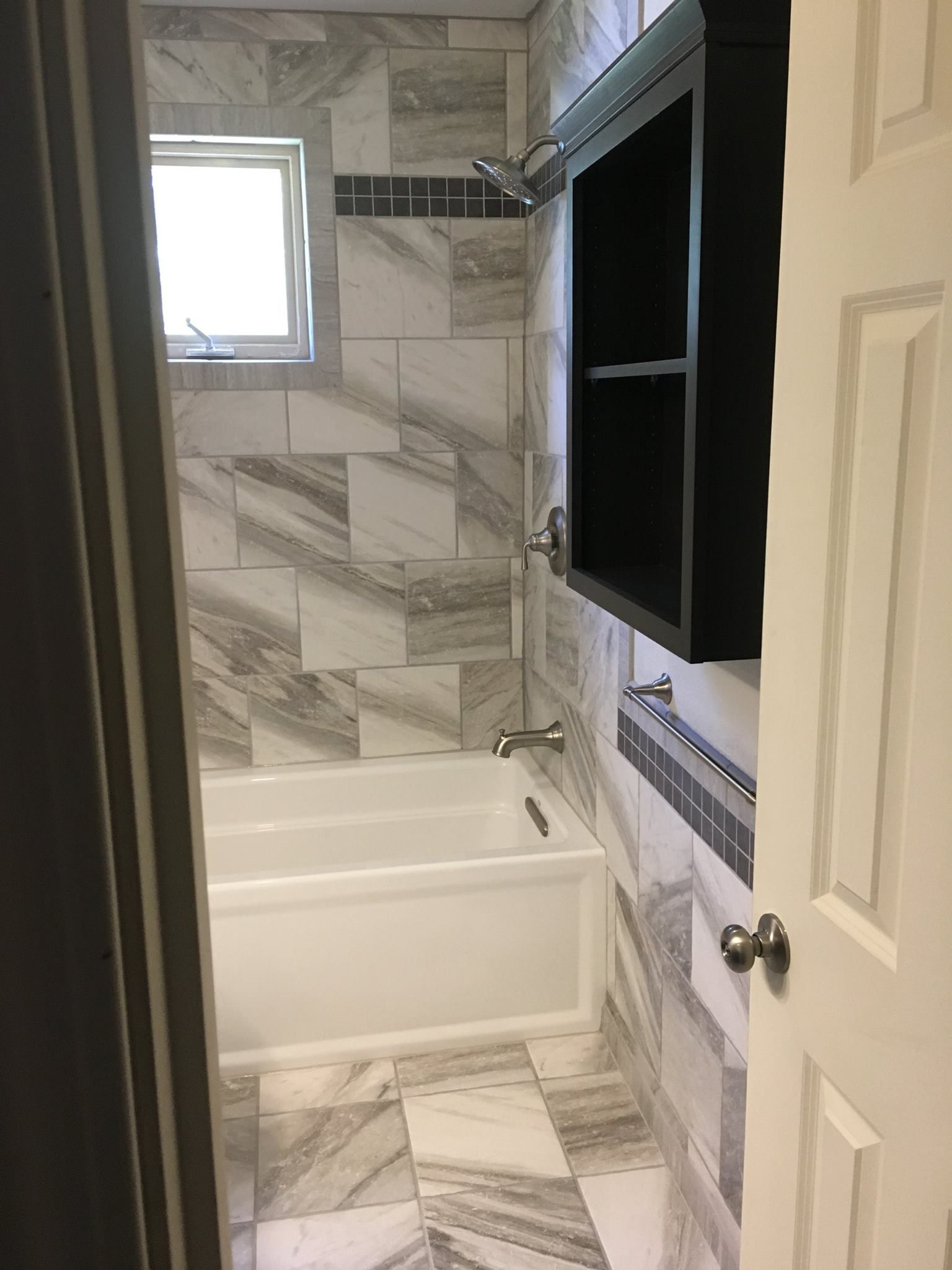 A bathroom with marble tile walls and floor. A white bathtub is in the middle, black cabinet on the right.