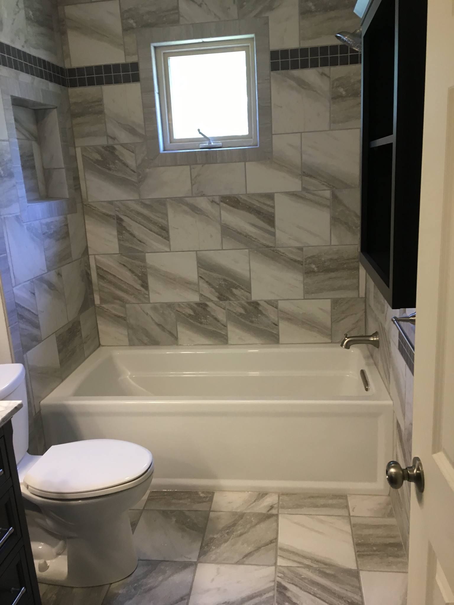 Bathroom with marble-patterned tile, white tub, toilet, and small window above.