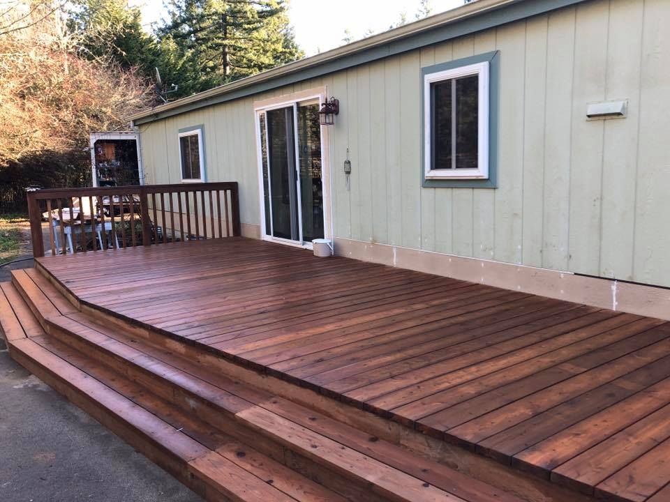 Newly stained wooden deck with steps attached to a light green house with a sliding glass door and windows.