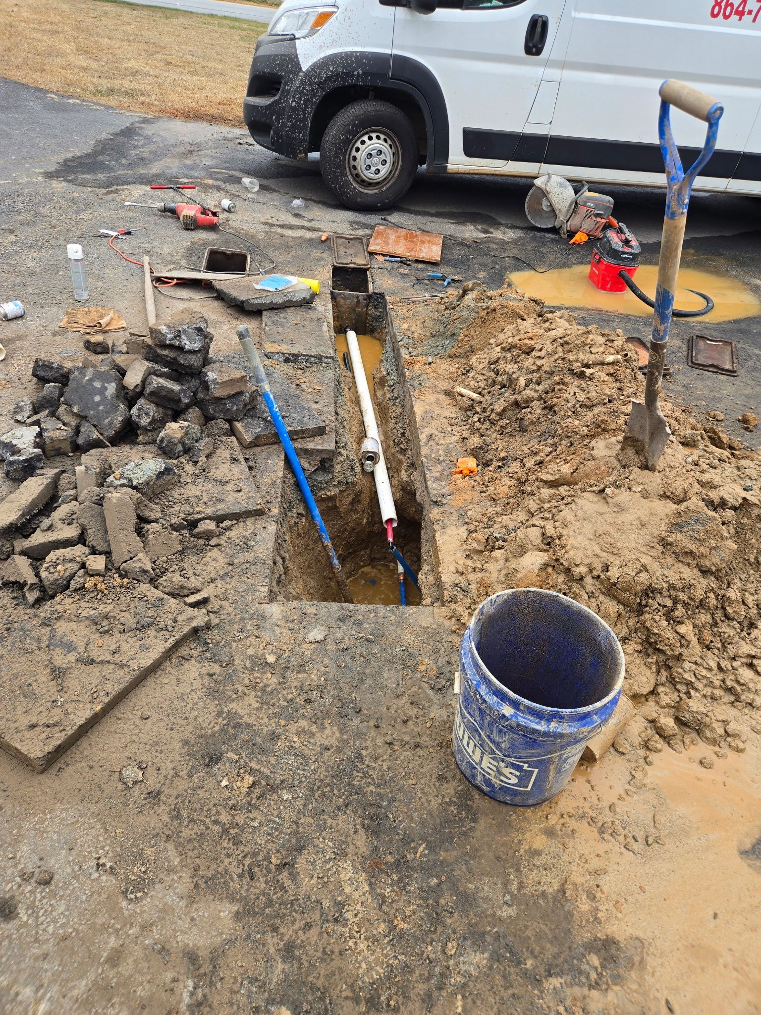 Trench dug in asphalt with exposed water pipe. White work van, tools, and bucket visible.