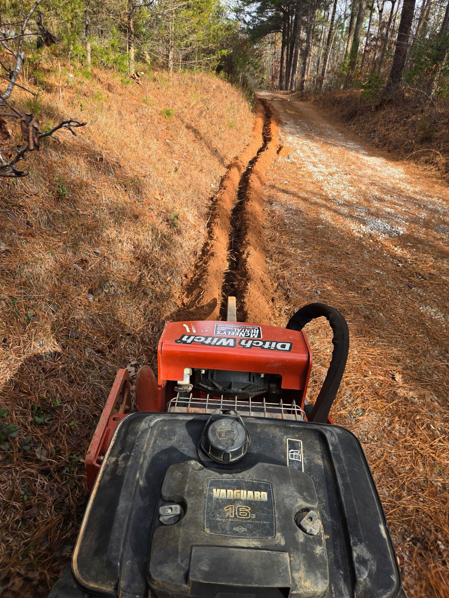 Trenching machine cutting a path through a dirt trail lined with leaves in a wooded area.