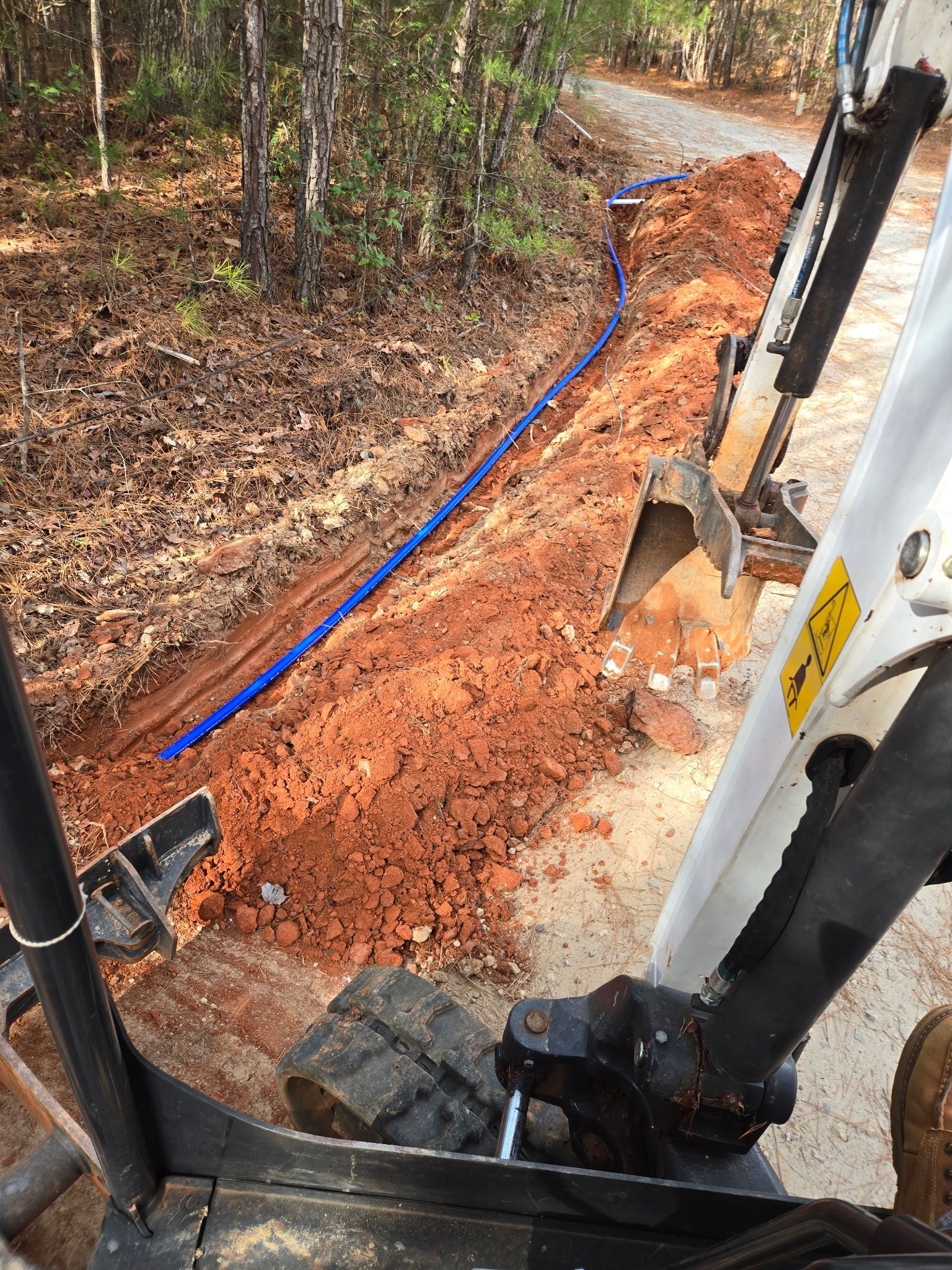 A blue pipe is laid in a trench dug by a small excavator on a dirt path, wooded background.