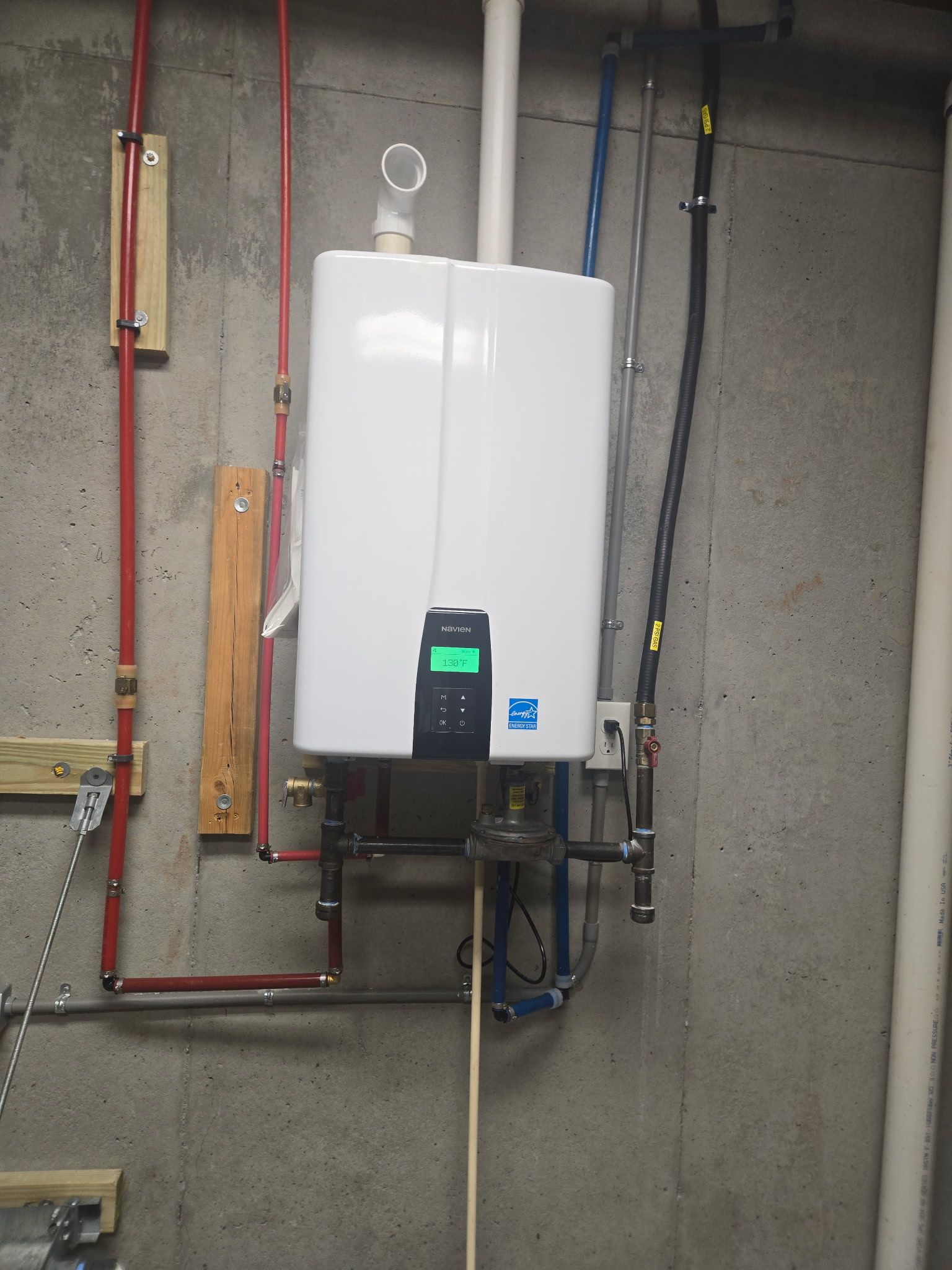 White wall-mounted water heater on a concrete wall with red and blue pipes.