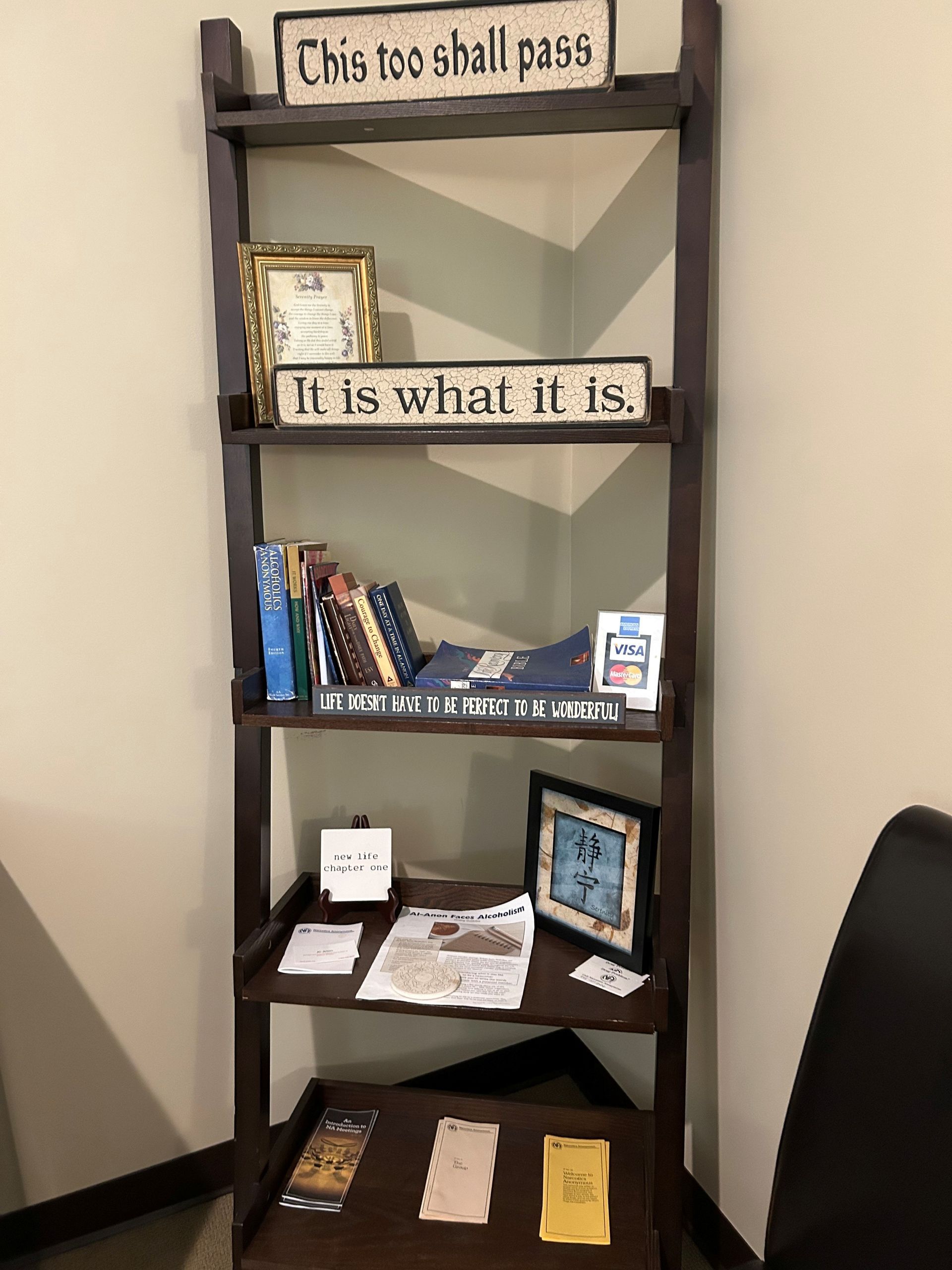 A bookshelf with a sign that says this too shall pass.