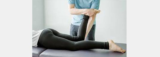 A person lies on a table while another person holds and stretches their leg; medical setting.