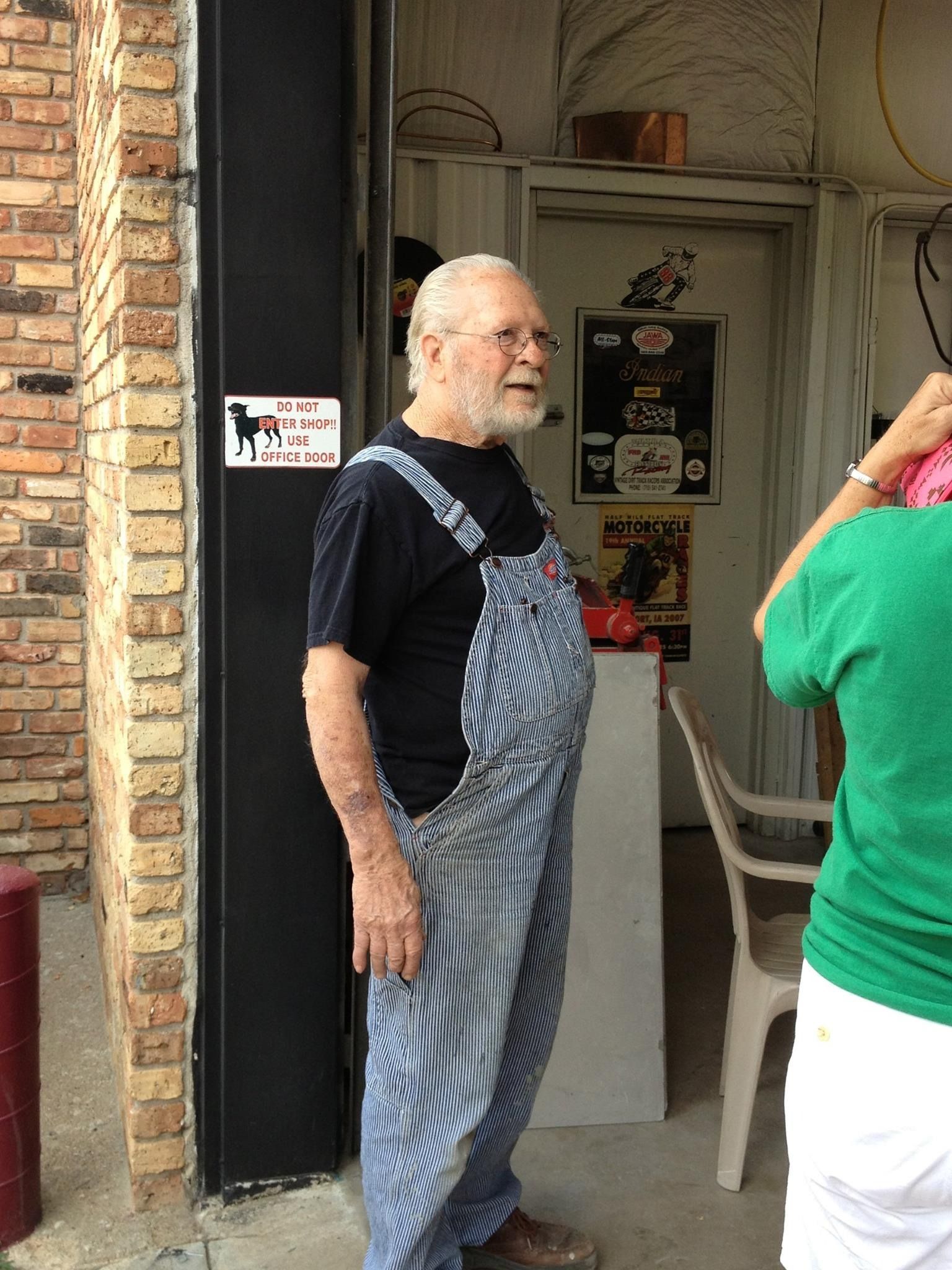 Man in blue striped overalls stands near a brick wall, talking to someone out of frame.