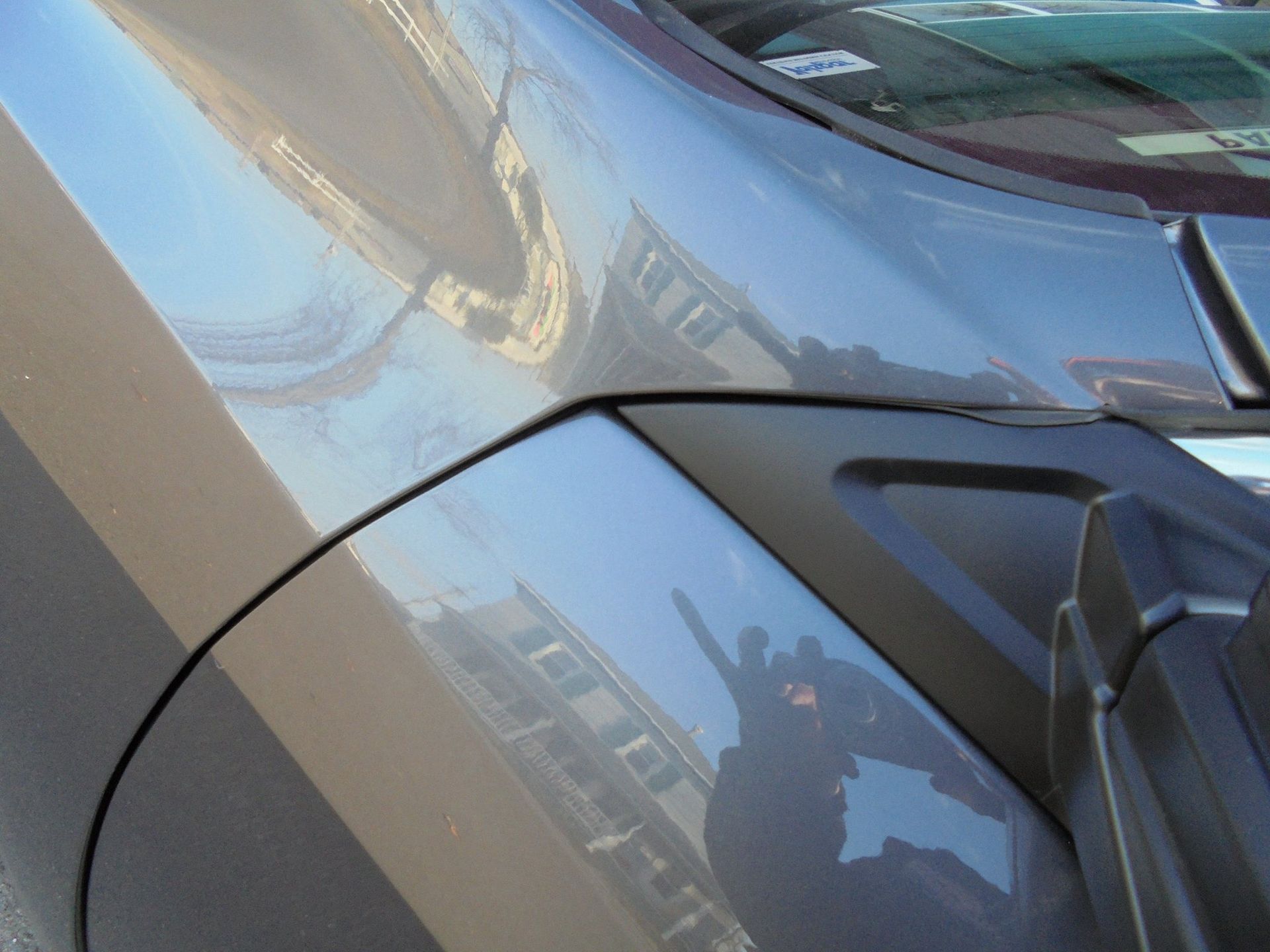 Close-up of a silver car's body, reflecting buildings and the sky, with a dark panel near the windshield.