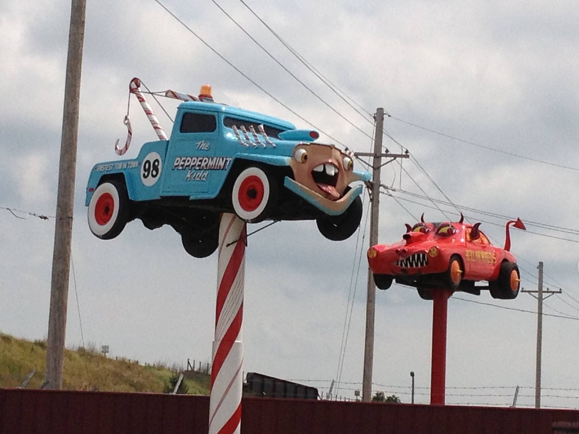 Large cartoon-like tow truck and race car sculptures on poles. Blue truck has teeth; red car has horns.
