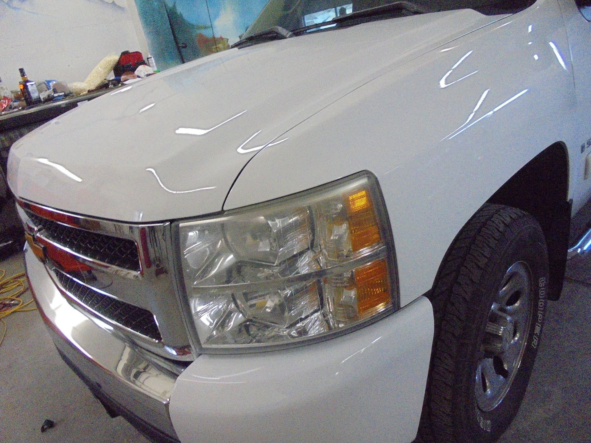 White Chevrolet truck's front end. Chrome grill, headlight, and bumper. Tire visible.