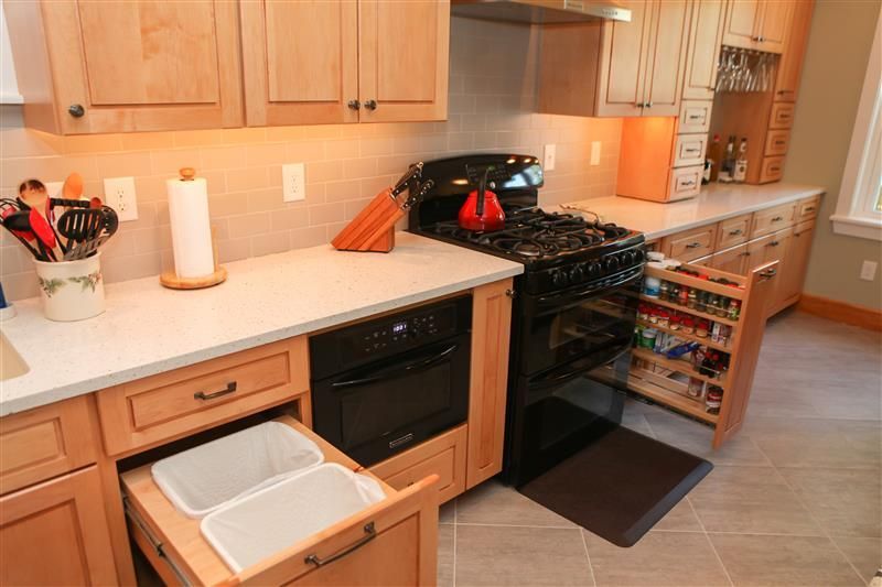 Kitchen with light wood cabinets, gray countertops, and a black stove. A trash drawer is open.