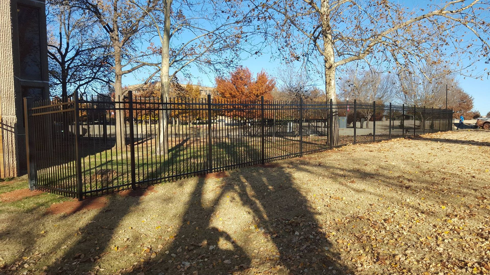 A metal fence surrounds a park with trees and leaves on the ground