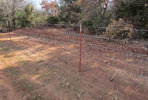 A barbed wire fence surrounds a dirt field with trees in the background.