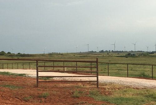 A fence surrounds a dirt road in a field with wind turbines in the background.
