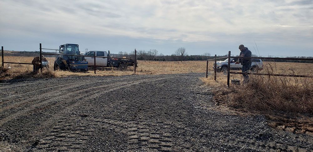 A man is standing in the middle of a dirt road next to a fence and a tractor