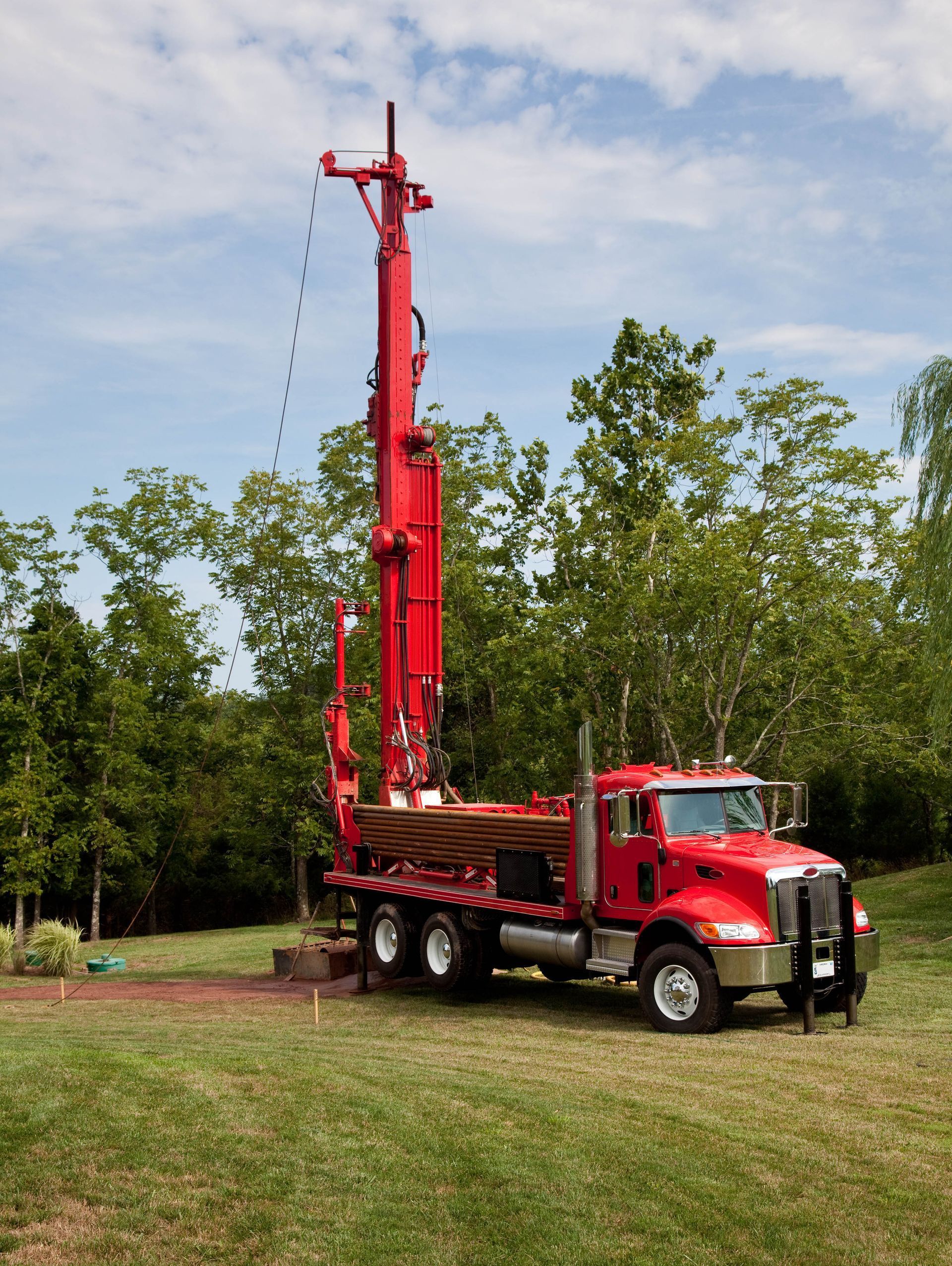 Red drilling rig on a truck in a grassy area, trees in the background, cloudy sky.