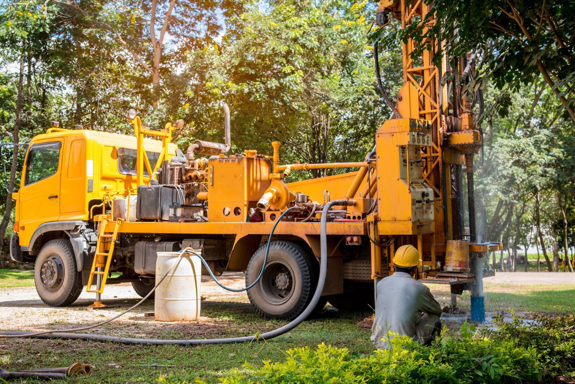 Yellow drilling rig on a truck, with a worker in a hard hat, operating near trees and a water hose.
