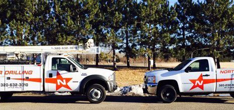 Two white work trucks with orange logos parked on a road. Trees and blue sky are in the background.