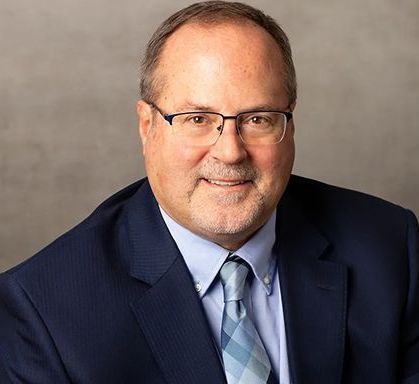 Man in a blue suit, glasses, and tie smiling, against a gray background.