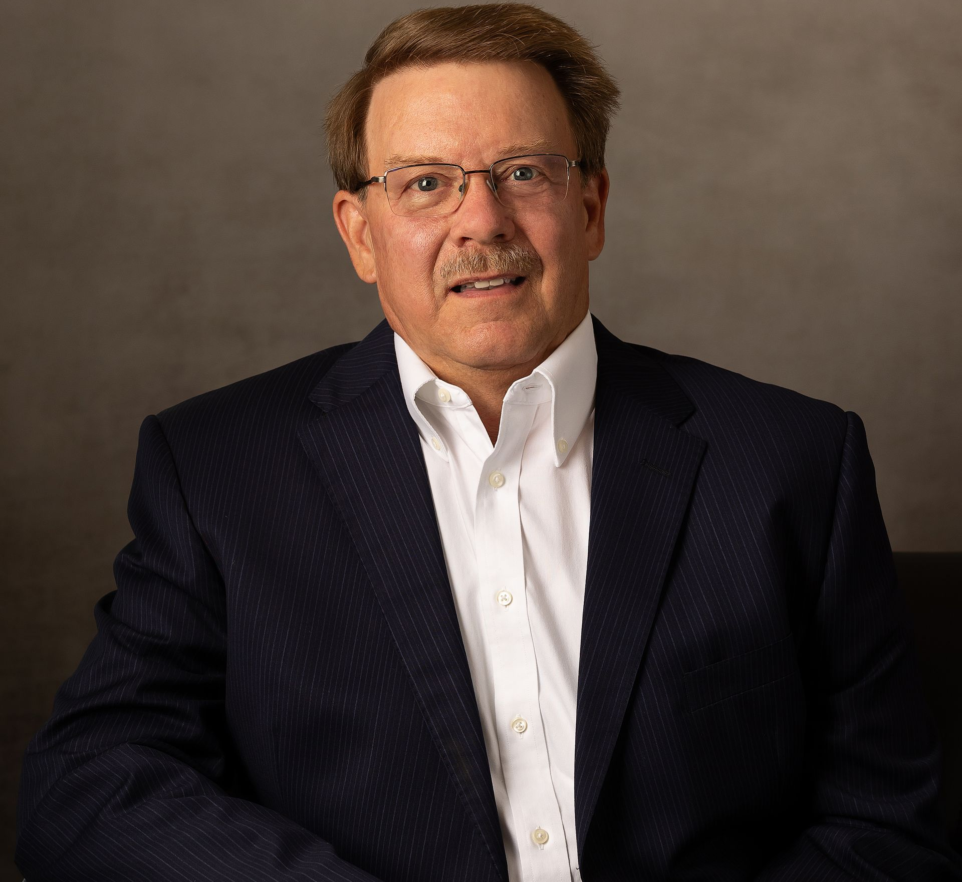 Man in suit jacket and glasses smiling, sitting against a neutral background.