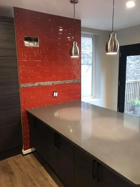 Kitchen with red tile accent wall, gray countertop, dark cabinets, and pendant lights.