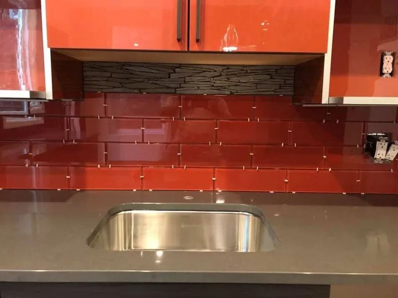 Red tile backsplash with stainless steel sink, under red cabinets, and gray countertop.