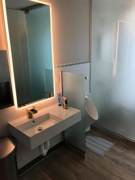 Modern bathroom with a sink, lit mirror, and a urinal behind a frosted glass partition.