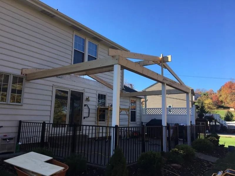 Construction of a wooden pergola attached to a white house, on a patio with a black fence.