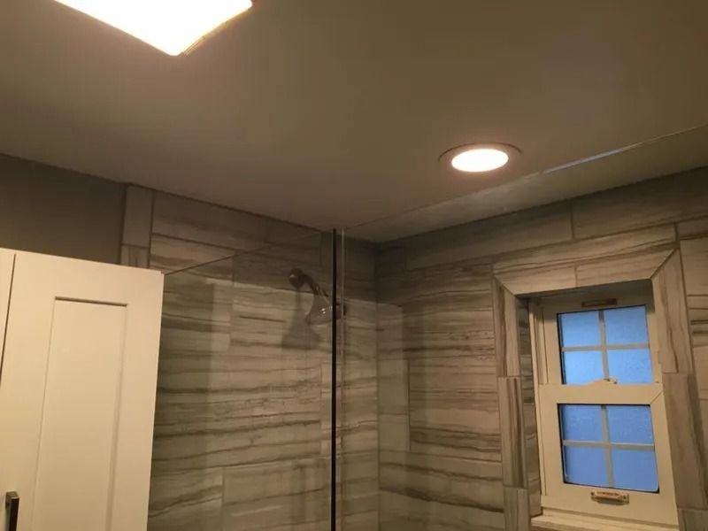 Bathroom interior with shower, window, and cabinetry. Gray tiled walls, white ceiling, and recessed lighting.