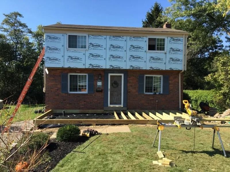 Two-story brick house under renovation with new deck framing. Blue insulation visible on upper level.