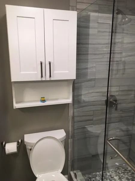 White bathroom cabinet above a toilet, with a glass shower enclosure and grey-tiled wall.