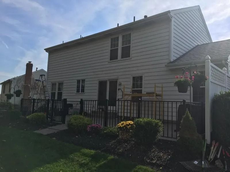 Back of a two-story beige house with a black fence, bushes, and a green lawn under a partly cloudy sky.