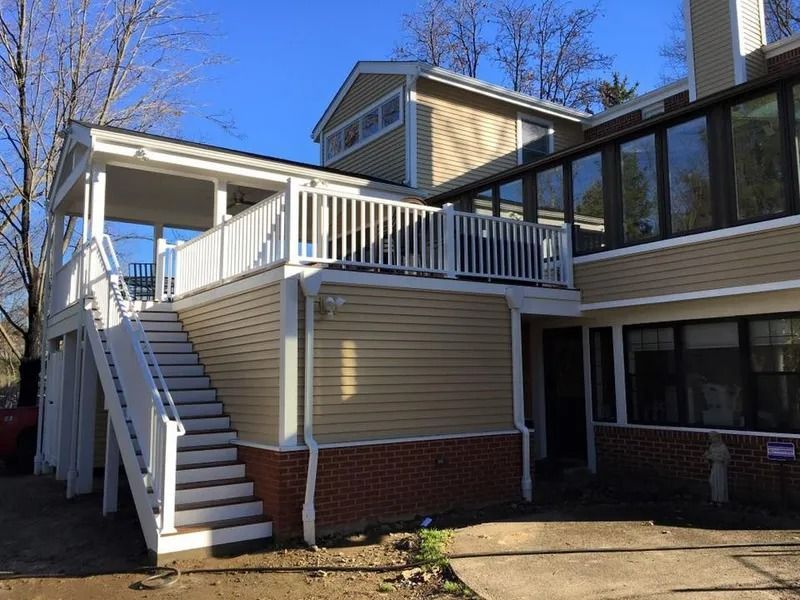 A two-story home with a deck and stairs. Tan siding with white railing and a red brick base.