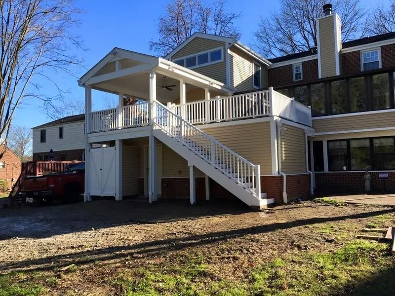 Two-story deck attached to a house with white railings, stairs, and light-yellow siding against a blue sky.