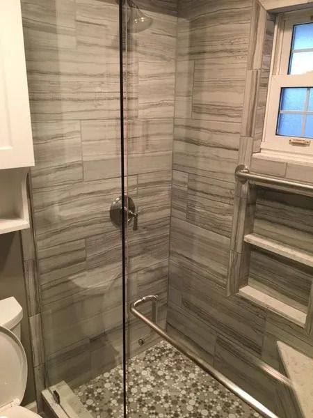 Bathroom shower with gray and white tiled walls, glass door, and pebble floor.
