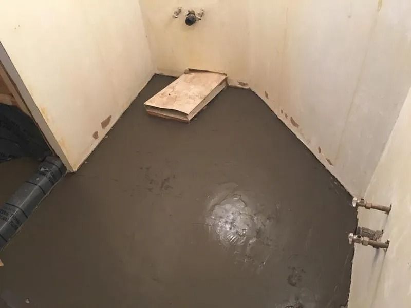 Bathroom with wet cement floor and a wooden board. Plumbing visible on the walls.
