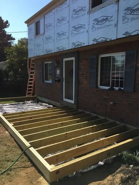 A house under construction with a deck frame.  Walls have siding wrap, brick, and windows.