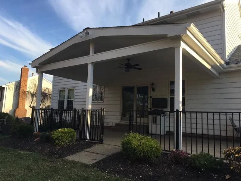 Covered patio with black railing, white columns, and a ceiling fan against a white house.