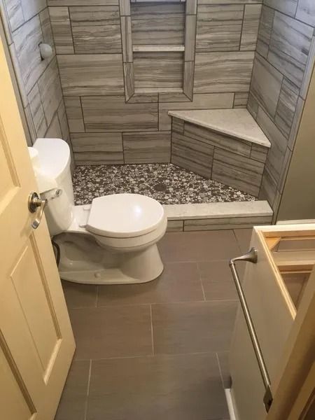 Bathroom with tiled shower, toilet, and gray floor tiles. The shower has a corner seat and a pebble floor.