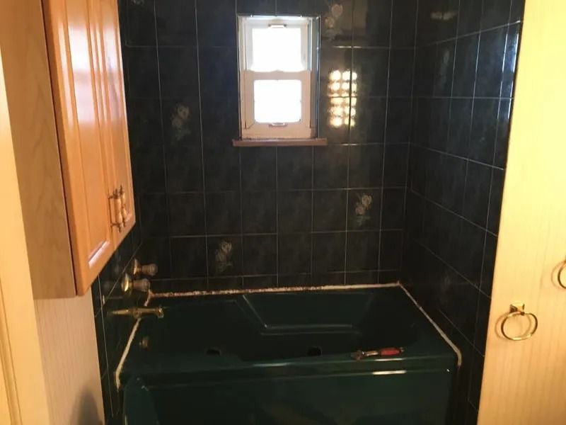 Bathroom with dark green tiled walls and tub, small window, and light wood cabinets.
