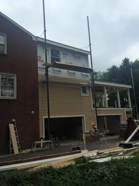 Construction of a two-story house addition with siding, a garage, and scaffolding. The sky is overcast.