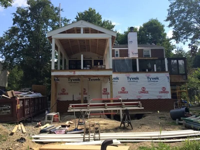 Home under construction with wooden framing, Tyvek wrap, and debris in the yard on a sunny day.