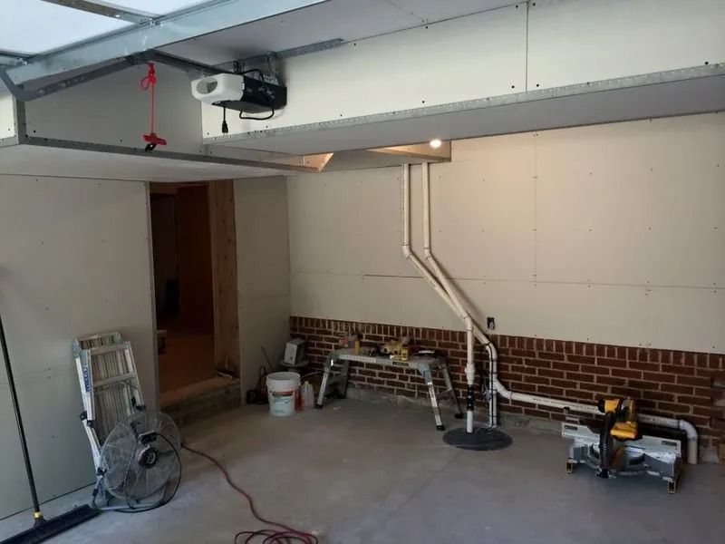 Garage interior with drywall and exposed pipes. Tools and fan on the floor. Doorway leads to another room.