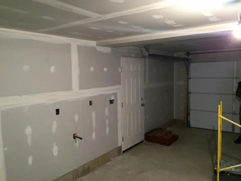 Garage interior with drywall installation in progress. Walls and ceiling are primed gray, door and garage door visible.