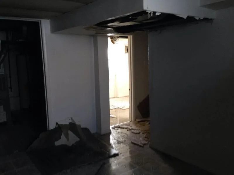 Interior view of a damaged room, showing exposed ceiling, debris on the floor, and a bright doorway.