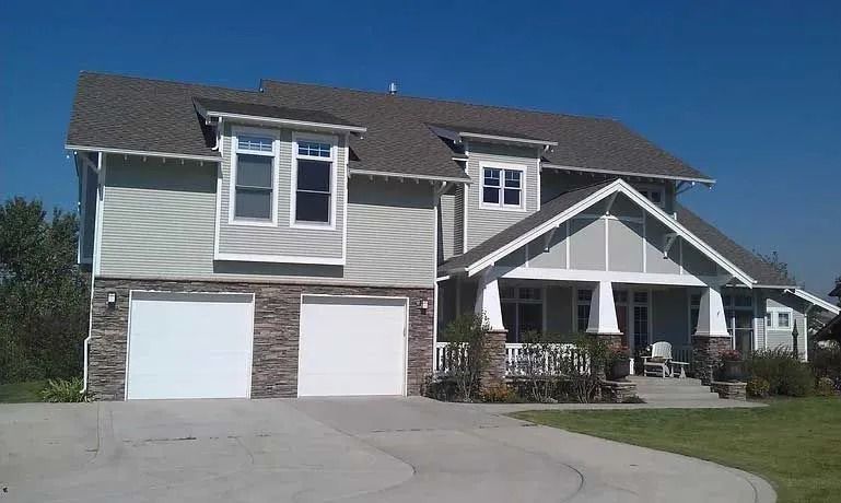 Two-story house with light green siding, stone accents, white garage doors, and a porch under a clear, blue sky.