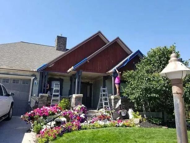 A house being painted; a person on a ladder is working on the red wood trim under a blue tarp.