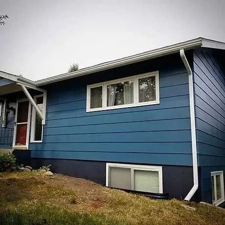 Blue house with white trim, windows, and gutters. Dark base on a sloped yard.