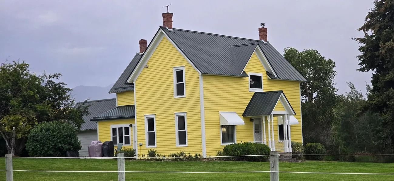 Yellow house with dark roof on a grassy lawn with trees in the background under a cloudy sky.