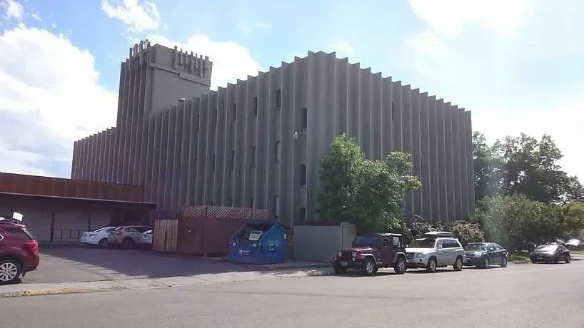Gray ribbed-sided building with square tower and parked vehicles in front. Trees and blue sky visible.