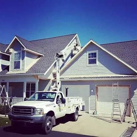 Workers on a lift paint the gable of a two-story house, a utility truck in front.